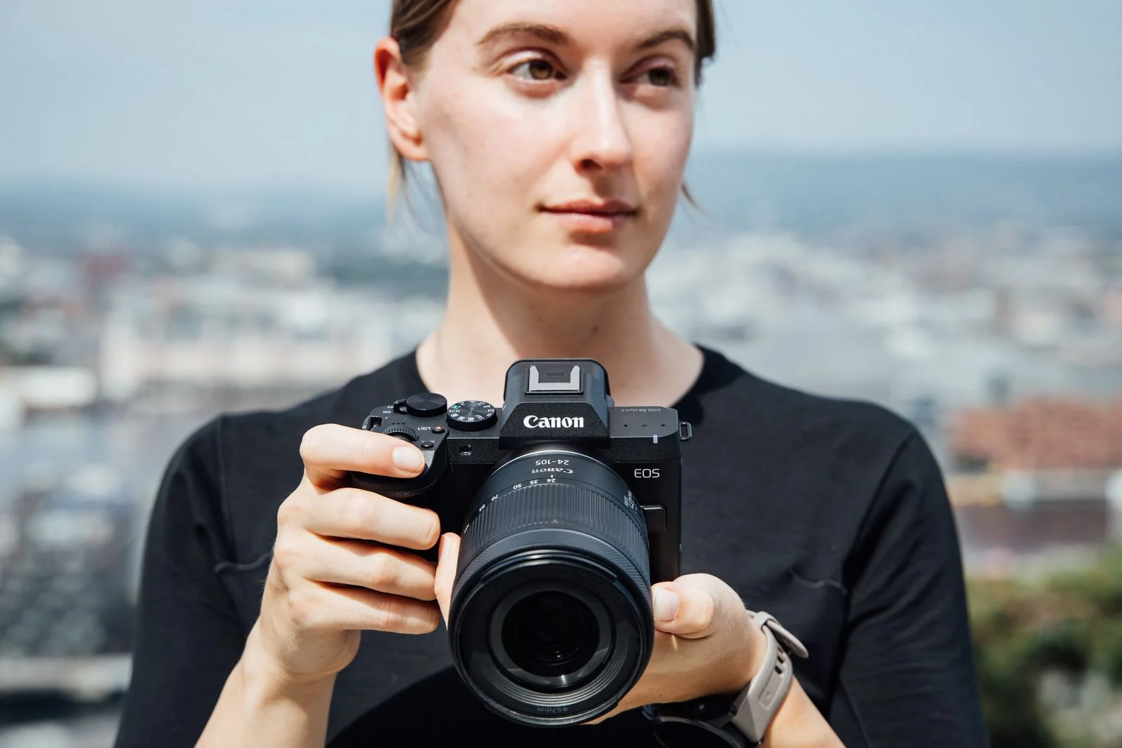 ผู้หญิงถือกล้อง Canon EOS R8 Mark II ไว้ระดับอก ฉากหลังเป็นเมือง