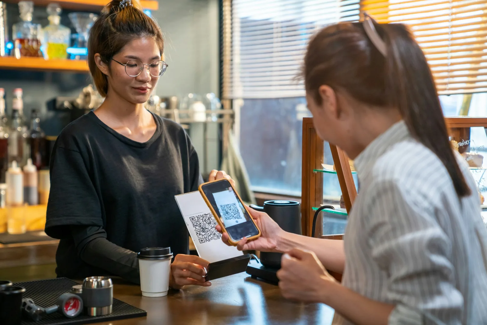 ลูกค้าสแกนคิวอาร์โค้ดจ่ายเงินที่เคาน์เตอร์ร้านกาแฟ พนักงานถือป้ายคิวอาร์