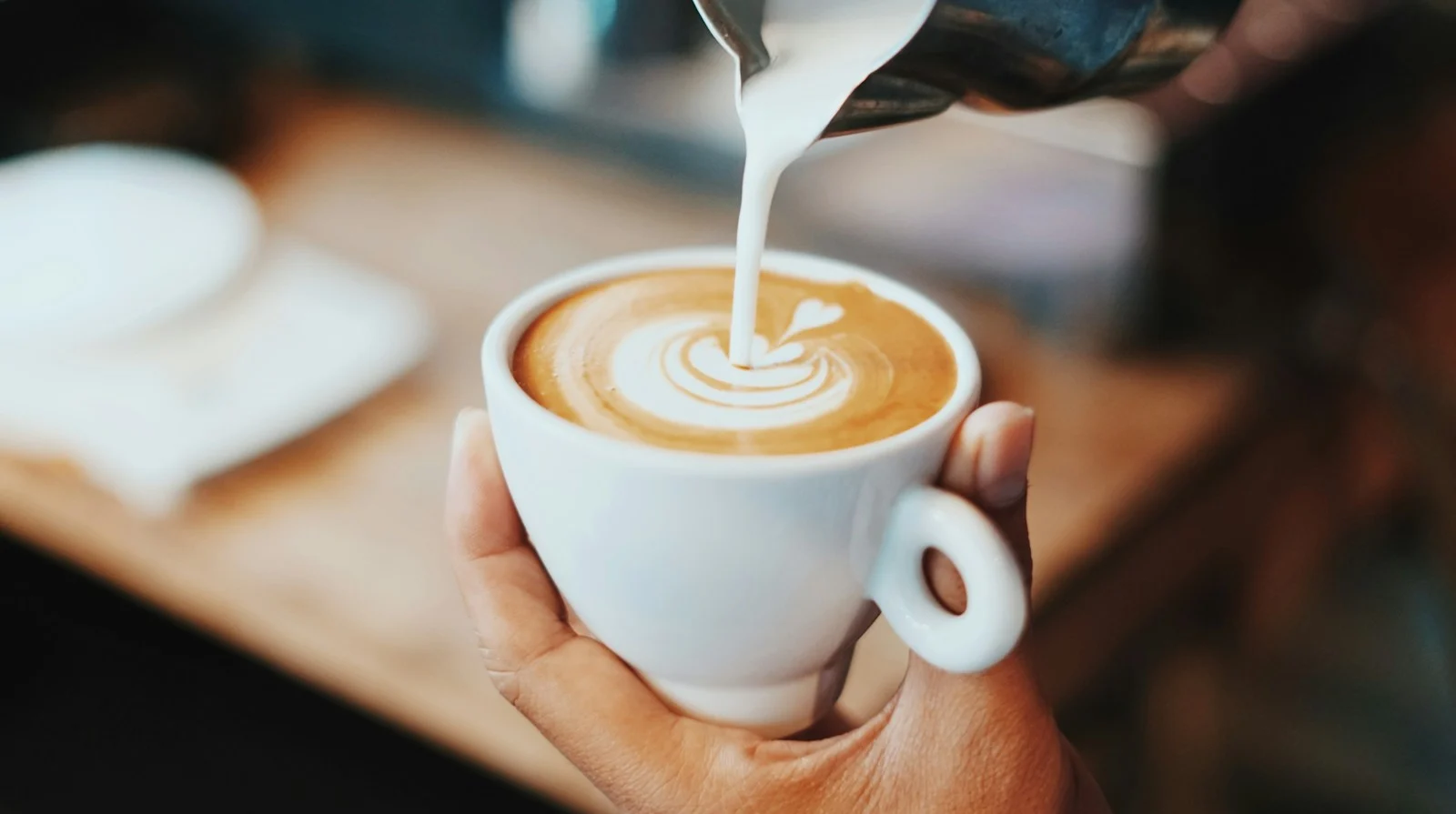 ภาพใกล้มือคนกำลังเทนมทำลาเต้อาร์ตลงในแก้วกาแฟ