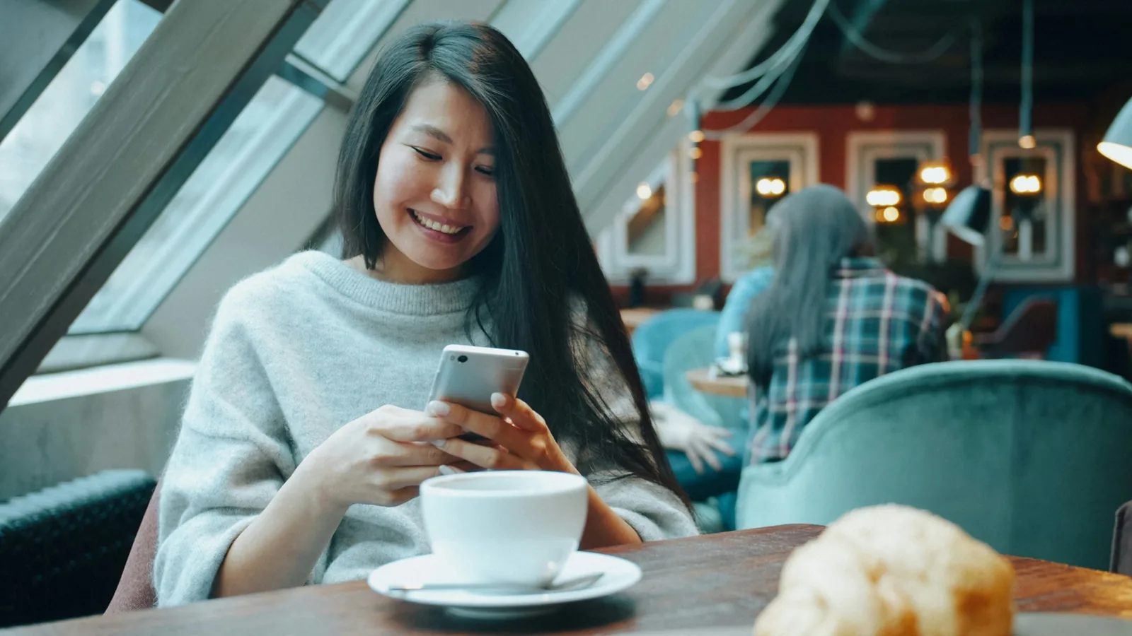 ผู้หญิงนั่งในคาเฟ่ ยิ้มขณะดูมือถือ มีถ้วยกาแฟวางบนโต๊ะ